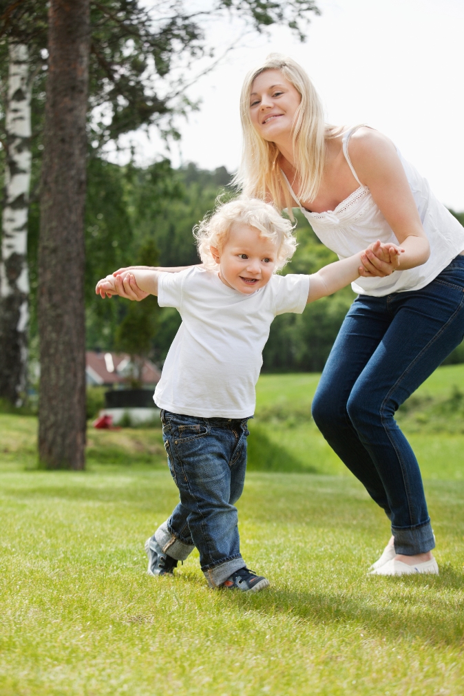 Beautiful young mother helping her adorable little boy to walk on grass