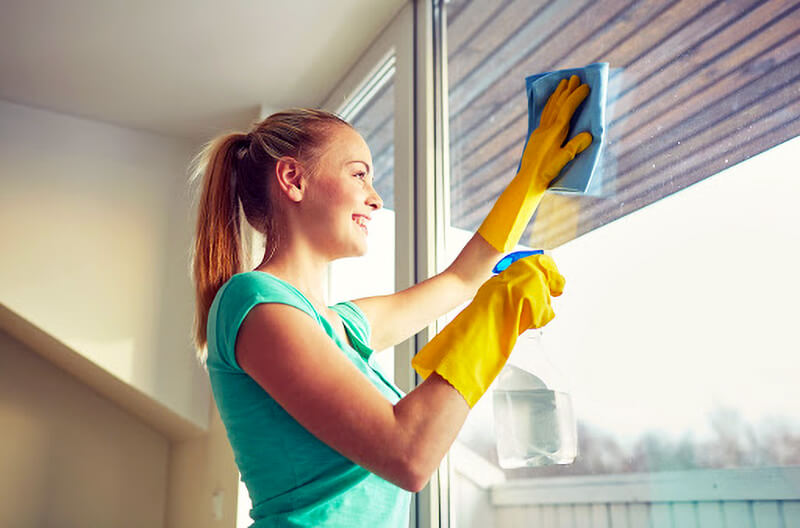 woman wearing yellow gloves smiling while cleaning windows with blue cloth